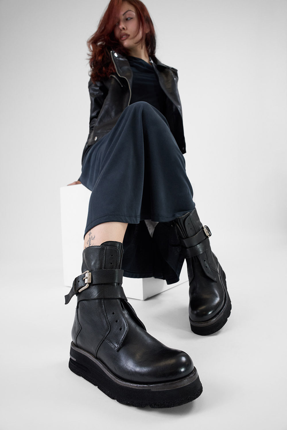 Person wearing black leather boots with a buckle in silver and a dark teal outfit on a white background